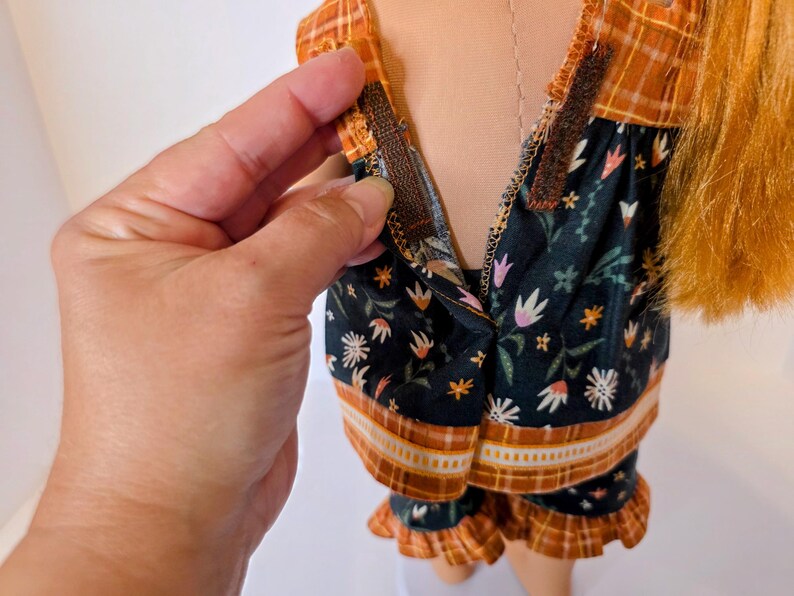May include: A doll wearing a two-piece pajama set. The top and shorts are dark green with a floral pattern and orange trim. The top has a plaid orange yoke. A hand is shown opening the zipper on the top.
