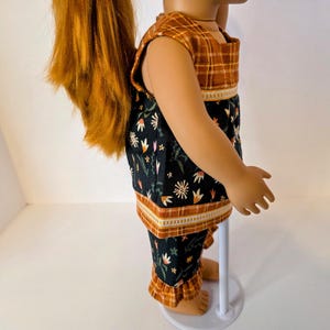 May include: A doll wearing a two-piece outfit. The top is sleeveless with a brown plaid pattern on the shoulders and a black floral print on the body. The matching pants have a floral print and ruffled hems.