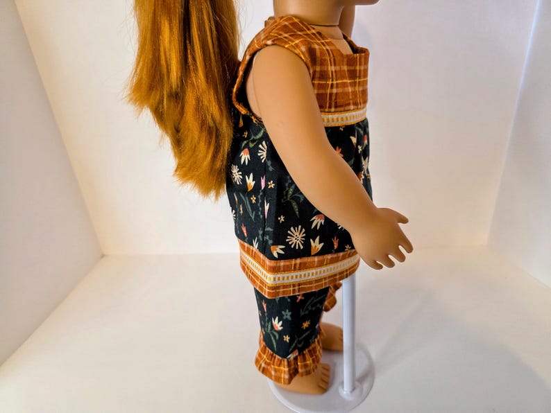 May include: A doll wearing a two-piece outfit. The top is sleeveless with a brown plaid pattern on the shoulders and a black floral print on the body. The matching pants have a floral print and ruffled hems.