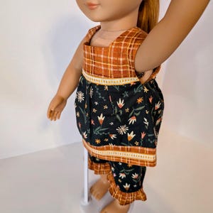 May include: Doll clothing set featuring a top and shorts. The top has a plaid yoke and a floral print body. The shorts have a matching floral print with a ruffled hem. The color palette includes orange, black, and white.