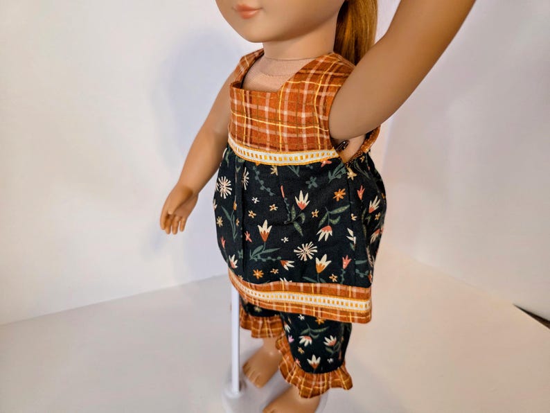 May include: Doll clothing set featuring a top and shorts. The top has a plaid yoke and a floral print body. The shorts have a matching floral print with a ruffled hem. The color palette includes orange, black, and white.
