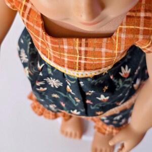May include: A doll wearing a two-piece outfit. The top is orange plaid, and the skirt is dark blue with a floral pattern. The outfit has a ruffled hem and a white trim. The doll's skin is light brown.