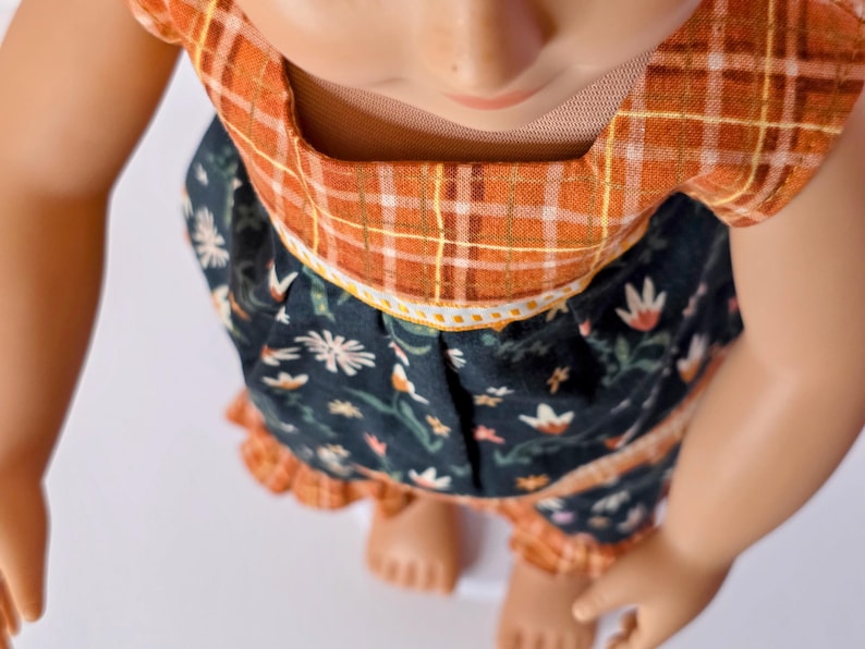 May include: A doll wearing a two-piece outfit. The top is orange plaid, and the skirt is dark blue with a floral pattern. The outfit has a ruffled hem and a white trim. The doll's skin is light brown.