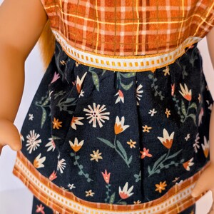 May include: A doll wearing a two-piece outfit. The top is a plaid pattern in orange and brown. The skirt is navy blue with a floral pattern in white, orange, and pink. The outfit has white and orange trim.