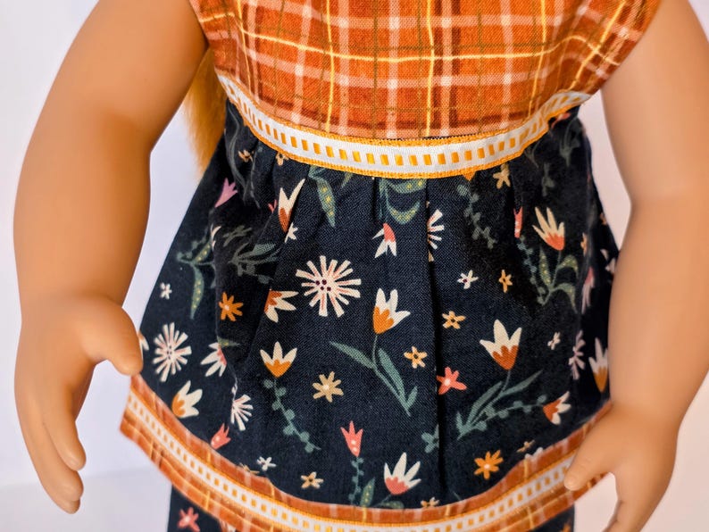 May include: A doll wearing a two-piece outfit. The top is a plaid pattern in orange and brown. The skirt is navy blue with a floral pattern in white, orange, and pink. The outfit has white and orange trim.