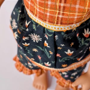 May include: A doll wearing a two-piece outfit. The top is orange plaid, and the skirt is dark blue with a floral pattern. The skirt has a ruffled hem. The outfit is designed for a doll.