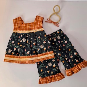 May include: A two-piece children's outfit featuring a dark floral print top and matching pants with ruffled hems. The top and pants have a plaid trim in shades of orange and brown. A matching headband with a bow is also included.