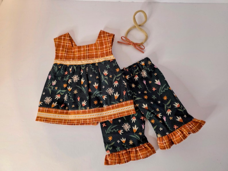 May include: A two-piece children's outfit featuring a dark floral print top and matching pants with ruffled hems. The top and pants have a plaid trim in shades of orange and brown. A matching headband with a bow is also included.