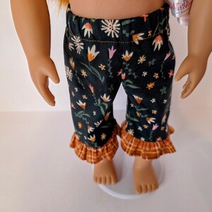 May include: Doll-sized pajama pants with a black background and a floral pattern in white, orange, and pink. The pants have a ruffled hem in a plaid pattern of orange, brown, and yellow.