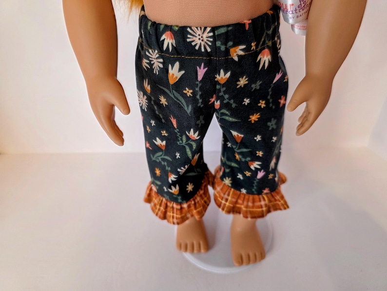 May include: Doll-sized pajama pants with a black background and a floral pattern in white, orange, and pink. The pants have a ruffled hem in a plaid pattern of orange, brown, and yellow.