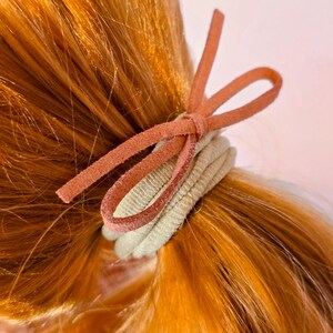 May include: A close-up of a ponytail secured with a light green hair tie and a rust-colored suede bow. The hair is a vibrant shade of auburn. The hair tie is made of a soft, ribbed fabric.