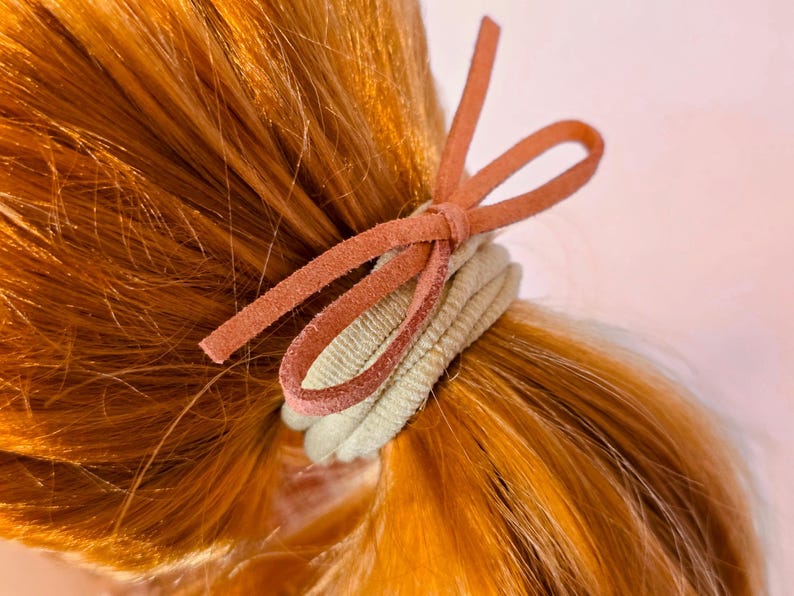 May include: A close-up of a ponytail secured with a light green hair tie and a rust-colored suede bow. The hair is a vibrant shade of auburn. The hair tie is made of a soft, ribbed fabric.