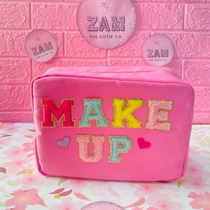 Könnte beinhalten: Eine rosa Schminktasche mit den Worten "MAKE UP" in bunten, flauschigen Buchstaben. Die Tasche hat einen Reißverschluss und ist mit mehreren rosa und violetten Kreisen verziert, auf denen "ZAM THE CUTIE CO." steht.