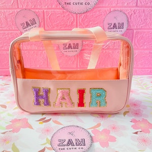 May include: A pink, clear cosmetic bag with the word "HAIR" in colorful, textured letters. The bag has a peach-colored interior and handles. The background features pink brick-like wallpaper and circular stickers with the text "ZAM THE CUTIE CO."
