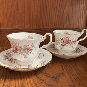 May include: Two white teacups and saucers with a floral rose pattern in pink and green. The teacups have gold trim and curved handles. The saucers have a scalloped edge and gold trim. The teacups and saucers are on a wooden surface.