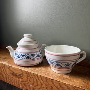May include: A white ceramic teapot with a lid and a matching teacup. Both pieces feature a blue and pink patterned design. The teapot has a spout and handle. The teacup has a handle. The set is placed on a wooden surface.