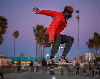 Peut inclure: Une personne portant une veste rouge et un pantalon noir effectue un trick de skateboard au-dessus d'un cône de signalisation. La personne porte des lunettes de soleil et a une planche à roulettes sous les pieds. L'arrière-plan est un skate park avec des palmiers et un ciel de coucher de soleil.