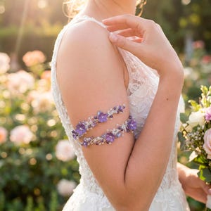 May include: A bride wears a white lace wedding dress and holds a bouquet of flowers. Two arm cuffs, adorned with purple flowers and crystals, encircle the upper arm. The background features a garden with soft sunlight.