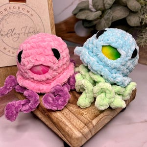 May include: Two handmade crochet octopus toys. One is pink with purple tentacles, the other is light blue with green tentacles. Both have black eyes and a colorful interior. They sit on a wooden block.