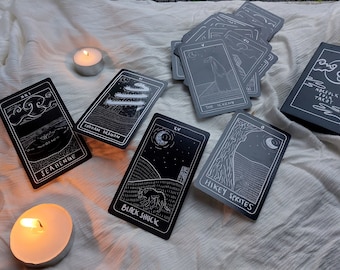 May include: A collection of black tarot cards with white illustrations and text, including "Seahenge" and "Black Shuck." The cards are arranged on a white cloth with lit tealight candles. A box with the text "Norfolk Folk Tarot" is also present.