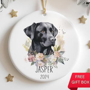 Op de afbeelding: Portret van een zwarte Labrador Retriever hond met een bloemenkrans en de naam "Jasper" en het jaar "2024" op een witte keramische hanger.