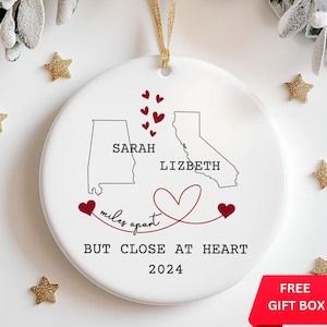 May include: White ceramic Christmas ornament with a red heart design and the text "miles apart but close at heart 2024". The ornament features outlines of Alabama and California with the names "Sarah" and "Lizbeth" written on each state.