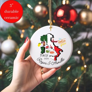May include: A white ceramic ornament with a map of Italy, featuring icons of Italian culture like a lemon, wine bottle, pizza, and a Vespa scooter. The text "Italy 2024 Roger & Armilla" and "La Dolce Vita" are printed on the ornament.