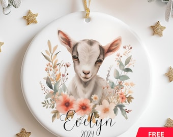 Könnte beinhalten: Weißer Keramikschmuck mit einer Aquarellillustration einer Ziege, umgeben von Blumen. Der Name "Evelyn" und das Jahr "2024" sind in Schrift geschrieben. Ein goldenes Band zum Aufhängen ist befestigt. Verziert mit goldenen Sternen.