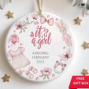 May include: A white ceramic ornament with a pink and white watercolor design. It features a baby girl's outfit, a bunny, a butterfly, a bow, and the text "it's a girl" and "arriving February 2025".