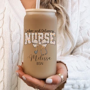 May include: A brown and beige reusable can-shaped tumbler with a lid and straw. The tumbler has a floral design with a stethoscope and bow. The text "Labor and Delivery Nurse" and "Melissa BSN" is printed on the tumbler.