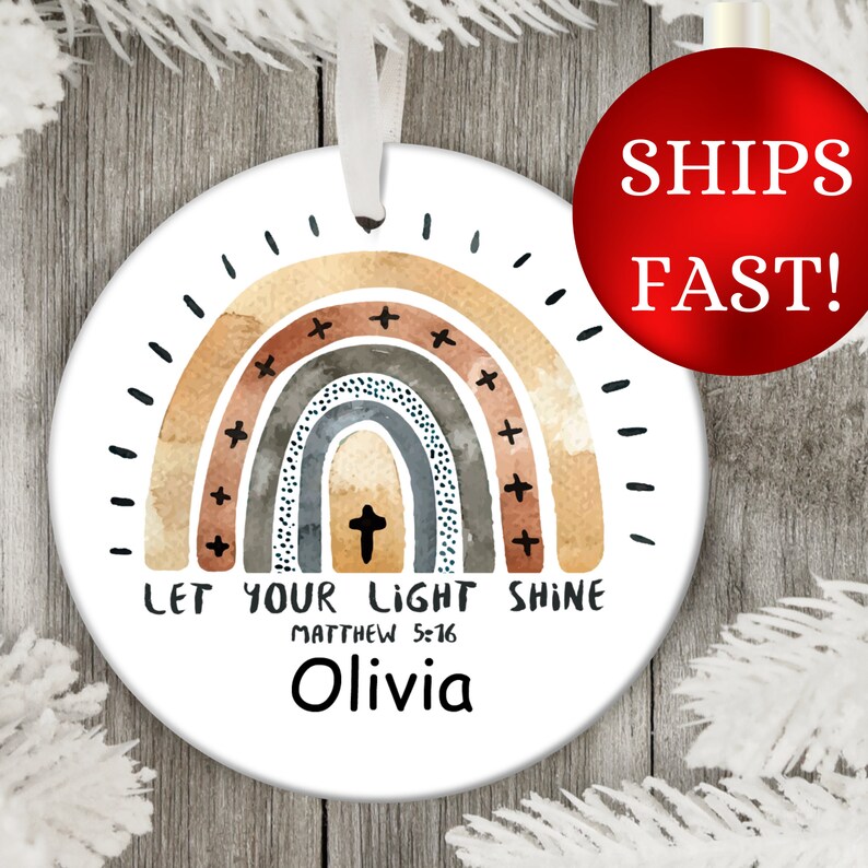 Puede incluir: Un adorno de cer&aacute;mica blanca con un dise&ntilde;o de arco&iacute;ris de acuarela en tonos marrones, grises y azules. El arco&iacute;ris tiene una cruz en el centro y el texto "Let your light shine Matthew 5:16 Olivia".