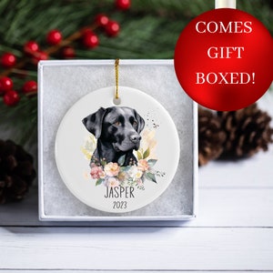 Op de afbeelding: Een ronde witte keramische kerstbal met een portret van een zwarte Labrador Retriever hond en bloemmotieven. De kerstbal is gepersonaliseerd met de naam "Jasper" en het jaar "2023".