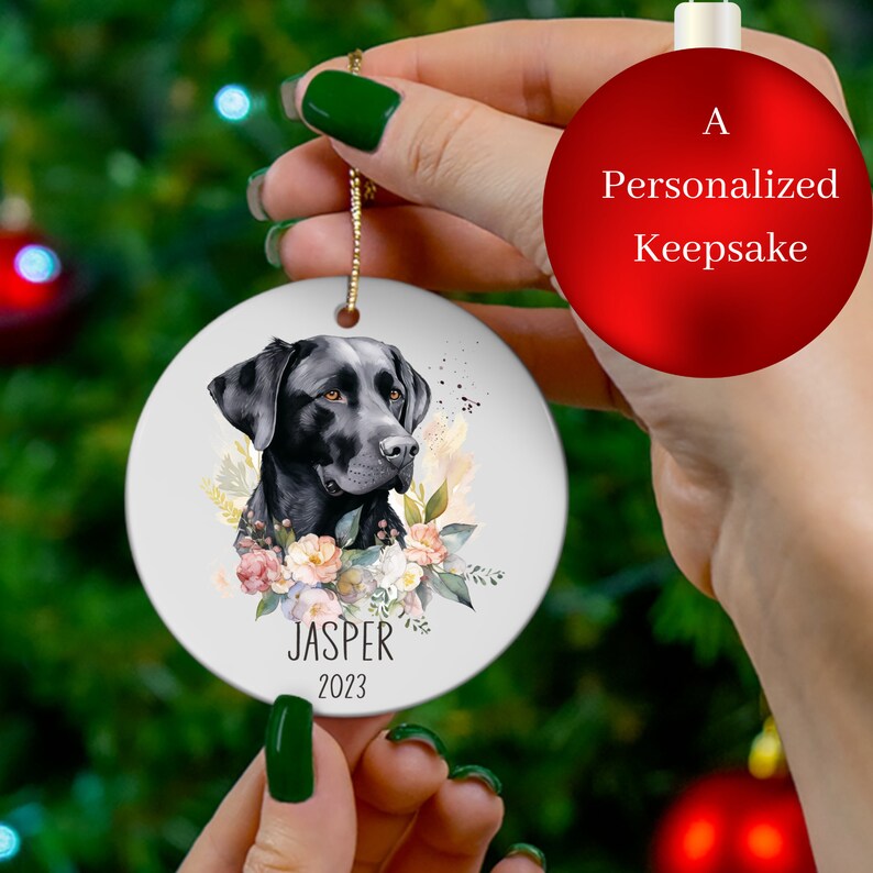 Op de afbeelding: Een witte keramische kerstbal met een portret van een zwarte Labrador Retriever, omringd door aquarelbloemen. De kerstbal is gepersonaliseerd met de naam "Jasper" en het jaar "2023".