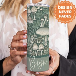 Puede incluir: Un vaso aislado verde y blanco con un dise&ntilde;o detallado de hongos y follaje. El vaso tiene una pajita y el nombre "Blythe" en una fuente de escritura. Tambi&eacute;n se ve el texto "DESIGN NEVER FADES".