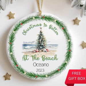 May include: White ceramic Christmas ornament with a green wreath border. The ornament features a watercolor illustration of a Christmas tree on a beach with the text "Christmas is Better At The Beach" and the year "2023".