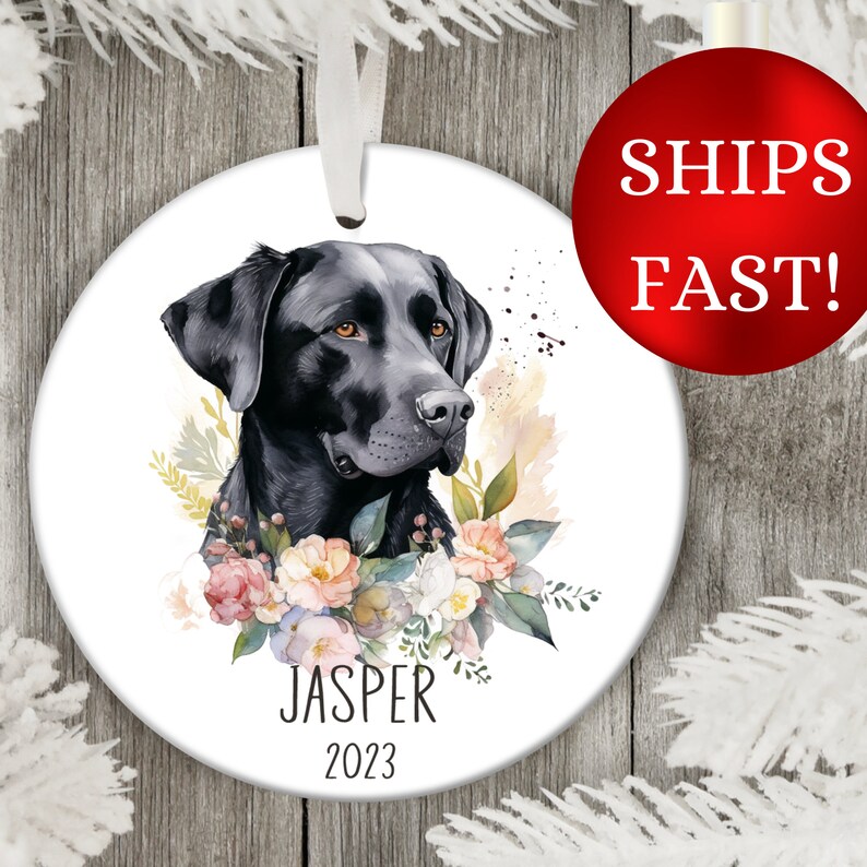 Op de afbeelding: Een ronde witte kerstbal met een zwarte Labrador Retriever met een bloemenkrans om zijn nek. De naam van de hond "Jasper" en het jaar "2023" staan onder de hond gedrukt.