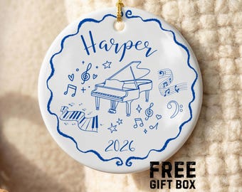 Puede incluir: Un adorno redondo de cerámica blanca con un borde azul festoneado. El diseño presenta una ilustración central de un piano de cola, notas musicales, estrellas y corazones en tinta azul. El nombre Harper está impreso arriba y el año 2026 abajo.