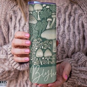 Puede incluir: Un vaso con tem&aacute;tica de setas verdes y blancas. El vaso presenta un patr&oacute;n repetido de varias setas y follaje. El nombre "Blythe" est&aacute; escrito en cursiva en la parte inferior. El vaso est&aacute; sostenido por una persona.