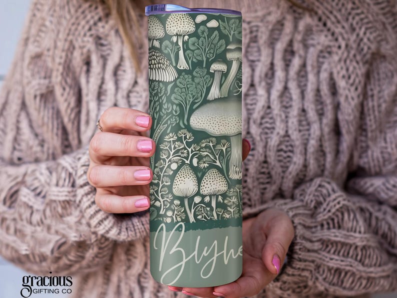 Puede incluir: Un vaso con tem&aacute;tica de setas verdes y blancas. El vaso presenta un patr&oacute;n repetido de varias setas y follaje. El nombre "Blythe" est&aacute; escrito en cursiva en la parte inferior. El vaso est&aacute; sostenido por una persona.