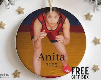 Puede incluir: Adorno redondo con una luchadora en un maillot rojo y negro sobre una colchoneta amarilla y roja. El nombre "Anita" y el año "2025" están impresos. También se ven estrellas doradas y el texto "FREE GIFT BOX".