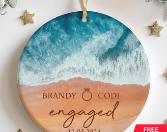 May include: Round, white ceramic ornament with a beach scene. The ornament features a blue ocean with white waves crashing on a sandy beach. The text "BRANDY & CODI engaged 12.03.2024" is printed on the ornament.