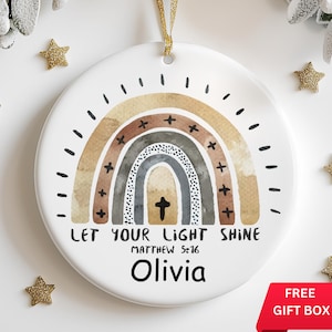 May include: A white ceramic ornament with a watercolor rainbow design in shades of brown, gray, and beige. The rainbow has a cross in the center and the text "Let your light shine Matthew 5:16" below it. The name "Olivia" is written in black at the bottom of the ornament.