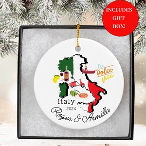 May include: White ceramic Christmas ornament with a map of Italy, featuring iconic symbols like a gondola, a Vespa scooter, lemons, olives, and a pizza. The text "Italy 2024 Roger & Armilla" and "La Dolce Vita" are printed on the ornament.