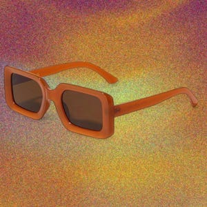 May include: Orange-tinted sunglasses with a rectangular frame and dark lenses. The sunglasses have a thick frame and arms, set against a glittery, multicolored background.