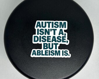May include: A black, circular sticker with the text "AUTISM ISN'T A DISEASE, BUT ABLEISM IS" in white and teal lettering. The sticker is centered on a black, textured surface.