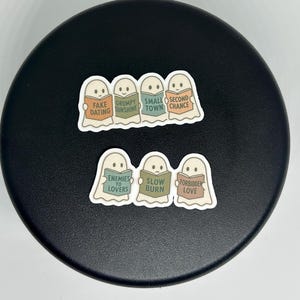 May include: A collection of stickers featuring ghost characters holding books. The books have titles such as "Fake Dating," "Grumpy Sunshine," and "Enemies to Lovers." The stickers are on a black surface.