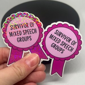 K&ouml;nnte beinhalten: Zwei rosa Aufkleber mit dem Text "SURVIVOR OF MIXED SPEECH GROUPS". Die Aufkleber haben die Form von Auszeichnungsb&auml;ndern mit einem gewellten Rand und einem holografischen Glitzereffekt. Ein Aufkleber wird in einer Hand gehalten.