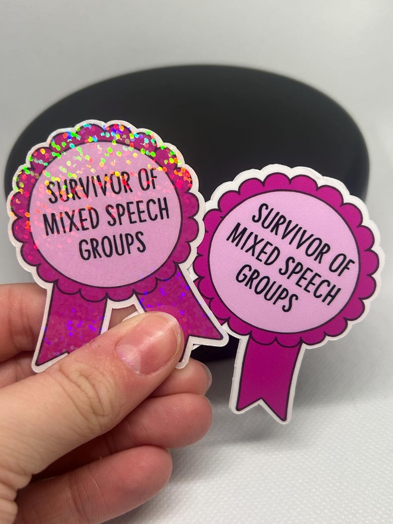 K&ouml;nnte beinhalten: Zwei rosa Aufkleber mit dem Text "SURVIVOR OF MIXED SPEECH GROUPS". Die Aufkleber haben die Form von Auszeichnungsb&auml;ndern mit einem gewellten Rand und einem holografischen Glitzereffekt. Ein Aufkleber wird in einer Hand gehalten.