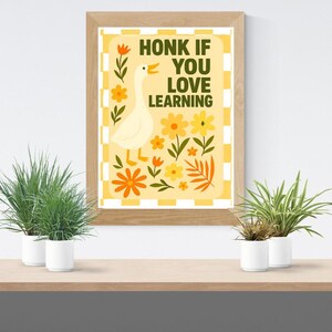May include: A framed print with the text "HONK IF YOU LOVE LEARNING" and a cartoon goose. The artwork features a yellow and orange color scheme with floral accents. The print is displayed on a wooden shelf with potted plants.