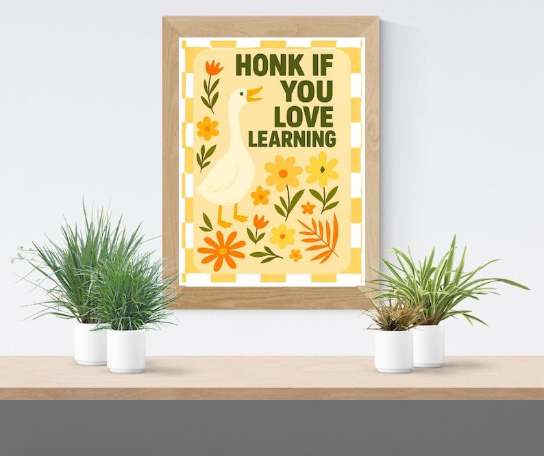 May include: A framed print with the text "HONK IF YOU LOVE LEARNING" and a cartoon goose. The artwork features a yellow and orange color scheme with floral accents. The print is displayed on a wooden shelf with potted plants.